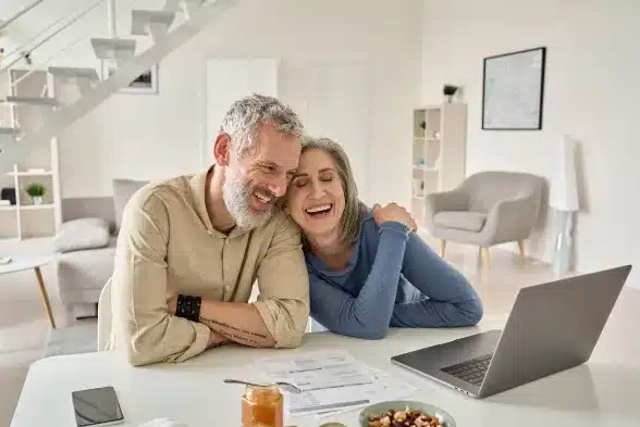 A happy couple seated at a table, engaged with a laptop, sharing a joyful moment together.