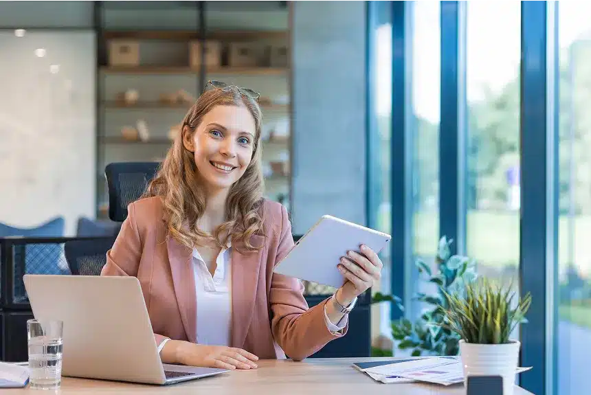 A woman sits at a desk, working on a laptop with a tablet beside her, focused on her tasks.