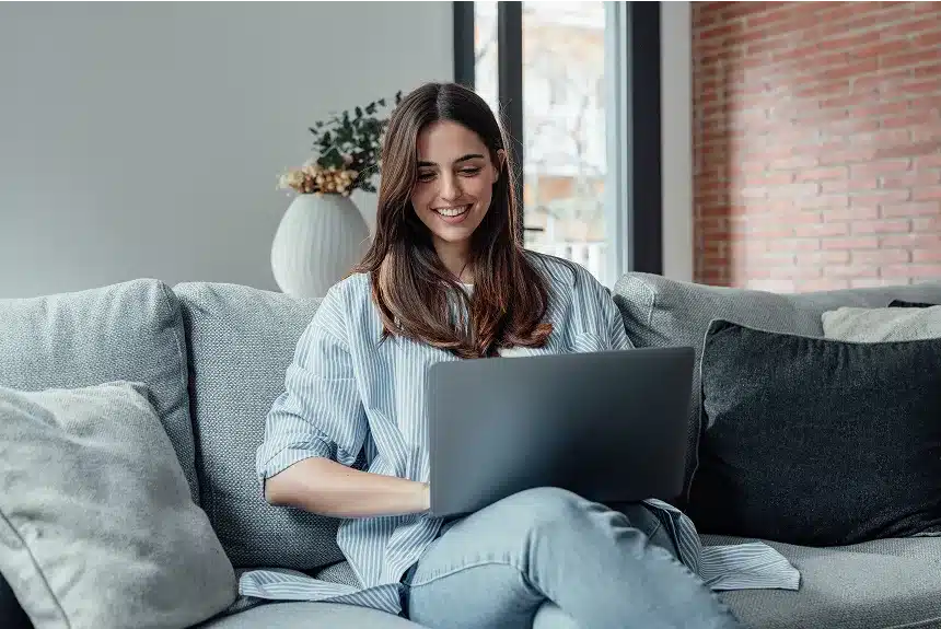 A woman sitting on a couch, focused on her laptop, with a cozy living room setting in the background.