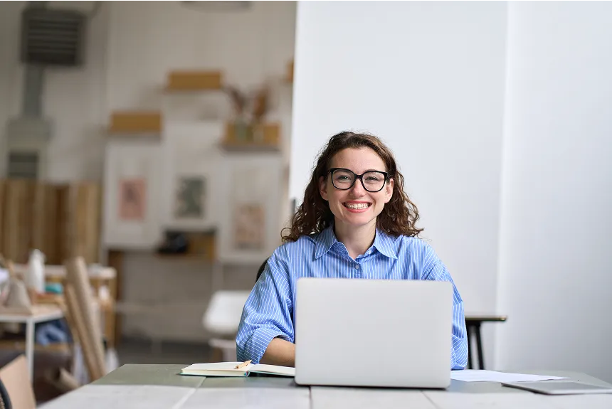 A woman wearing glasses sits at a table, focused on her laptop while working or studying.
