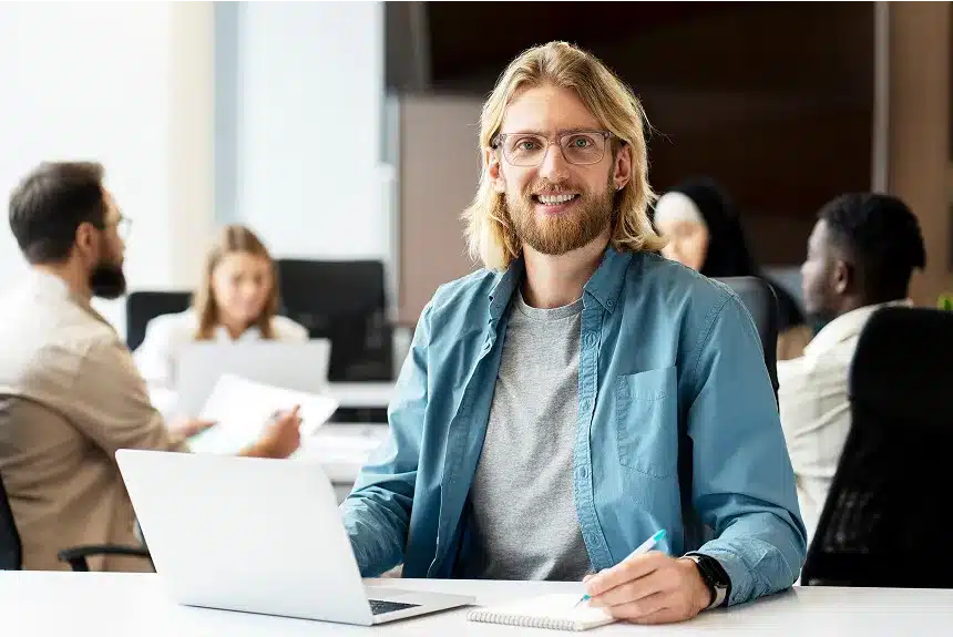 A man with glasses and a beard sits at a table, working on a laptop.