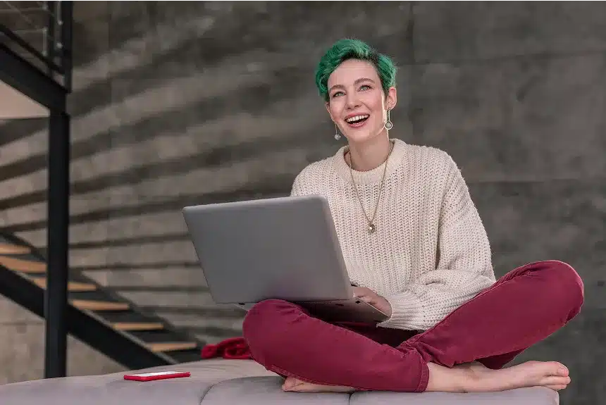 A woman with green hair sits on a couch, working on a laptop.