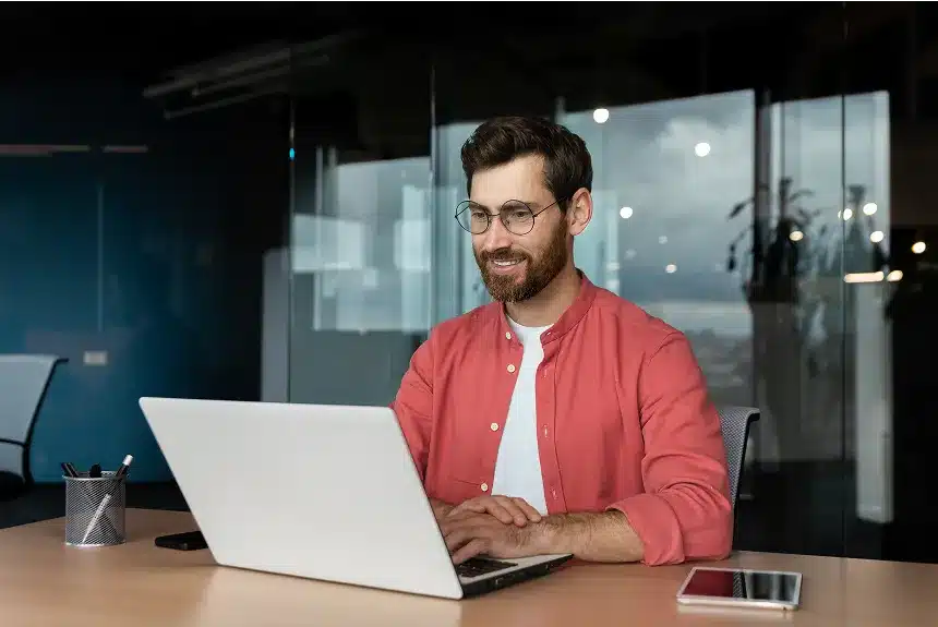 A man wearing glasses sits at a desk, focused on his laptop, with a neutral background.