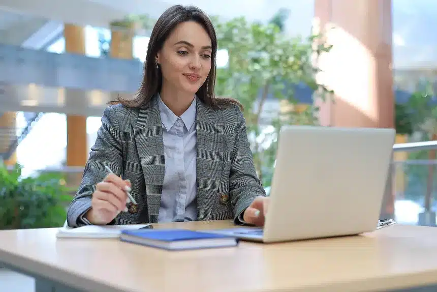 A woman in a business suit sits at a desk, focused on her laptop, with a professional and organized workspace around her.