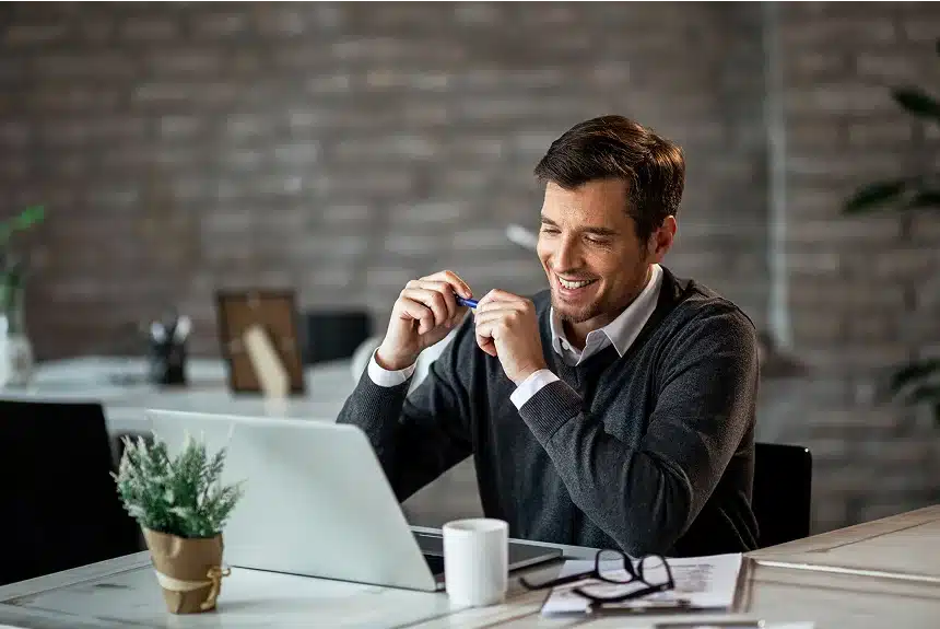 Smiling man in a sweater sits at a desk, looking at a laptop. He holds a pen, with papers, glasses, and a potted plant nearby, creating a cozy work atmosphere.