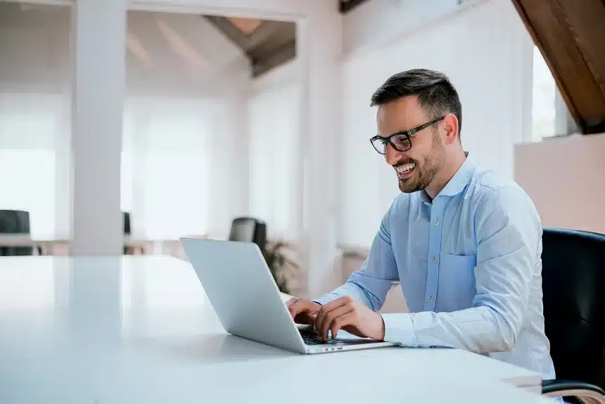 A man in glasses, wearing a light blue shirt, smiles while typing on a laptop in a bright, modern office, conveying a cheerful, productive atmosphere.