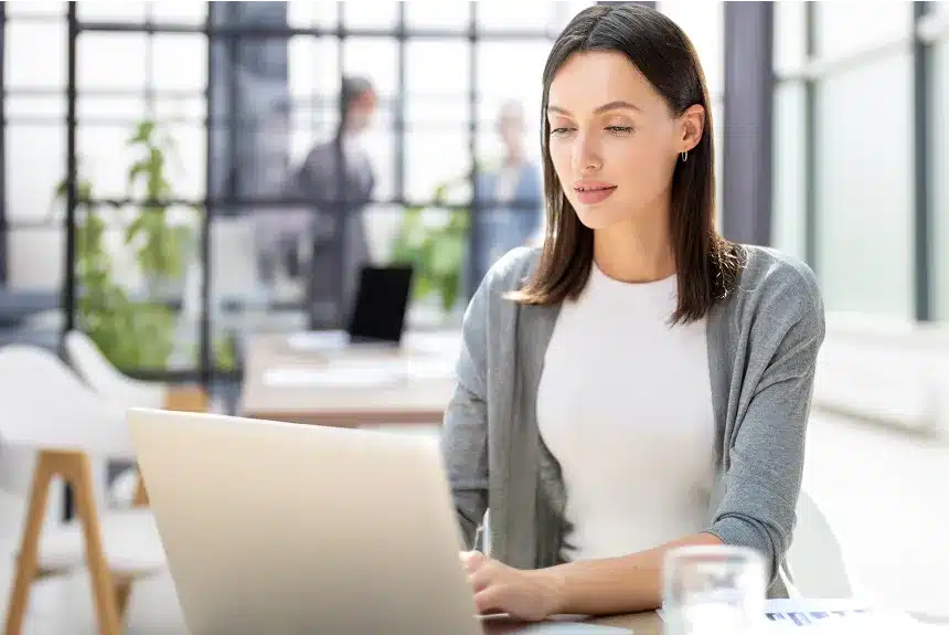 A woman sitting at a table, focused on her laptop, with a cozy indoor setting in the background.
