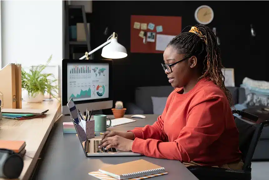 A woman in a red shirt sits at a desk, focused on her laptop while working.