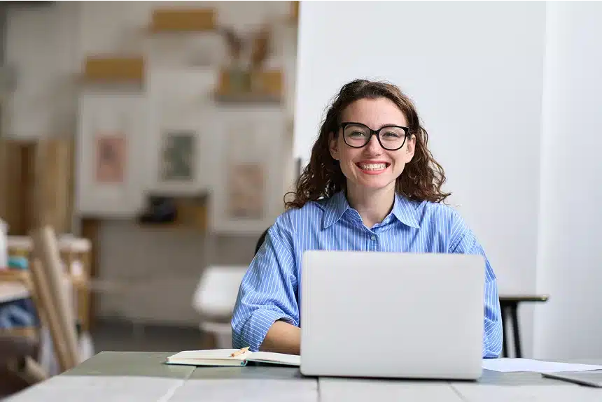 A woman with curly hair and glasses smiles while seated at a table with a laptop and notebook. She wears a blue striped shirt. The room has a creative, cozy vibe.