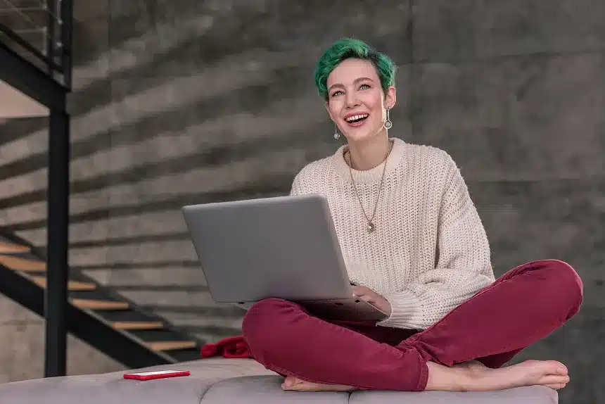 A woman with green hair sits on a couch, working on a laptop, surrounded by a cozy living room setting.