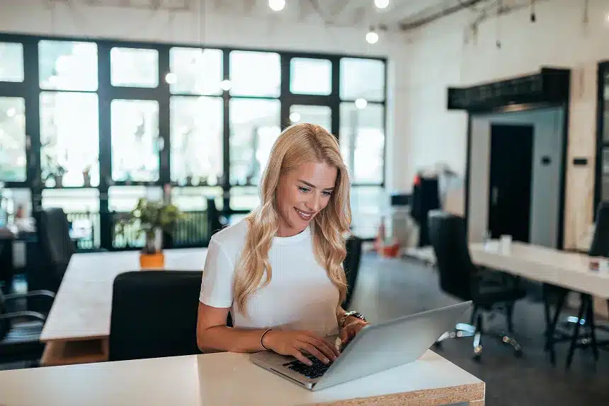 A woman sits at a table, focused on her laptop, with a calm expression in a well-lit indoor setting.