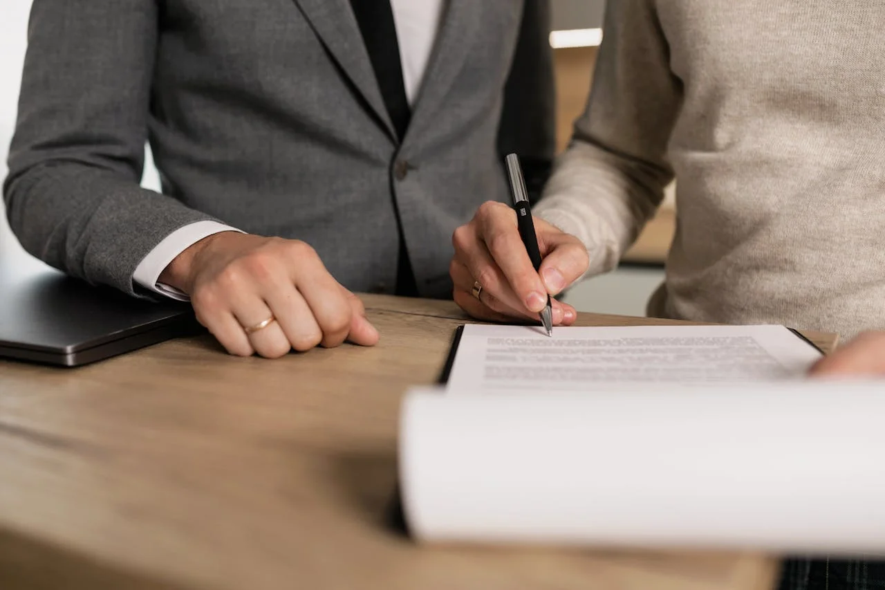 Two professionals in business attire signing documents at a table, indicating a formal agreement or contract.