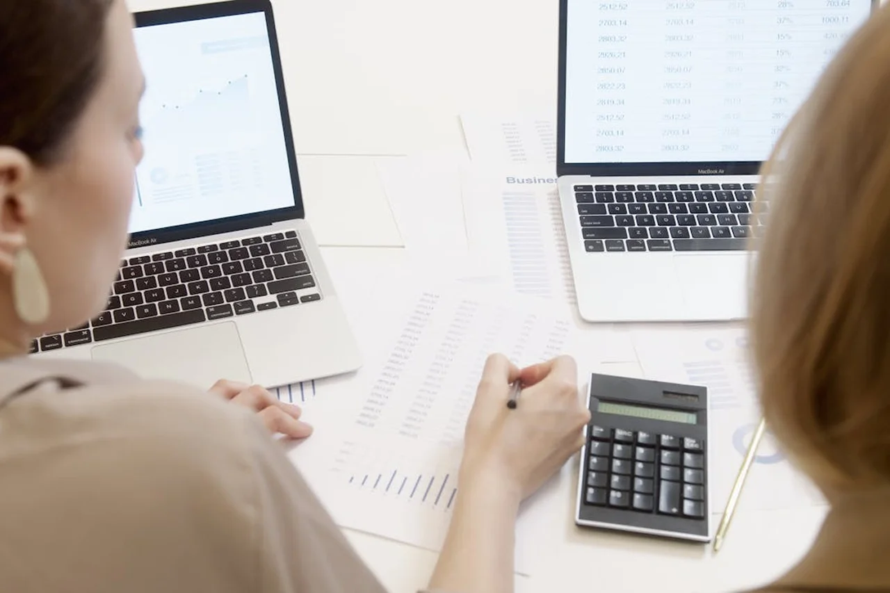Two women collaborate on a laptop while using a calculator for calculations.