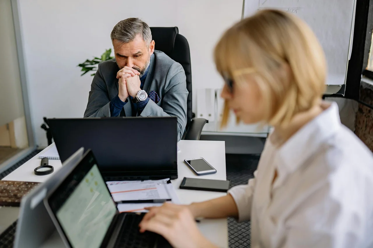 A man and woman sit at a desk, each using a laptop, engaged in a collaborative work session.