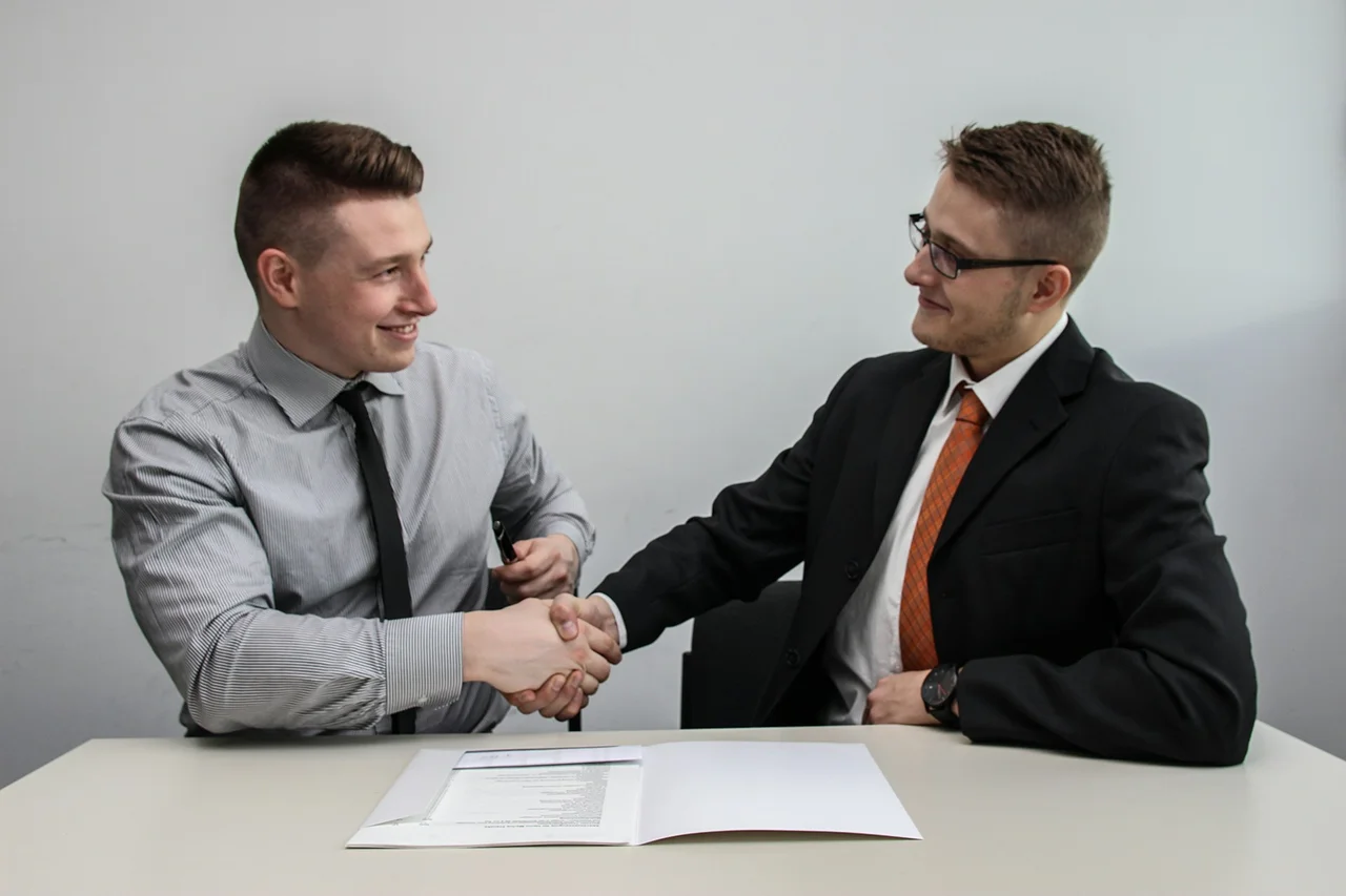 Two men shaking hands across a desk, symbolizing agreement or partnership in a professional setting.